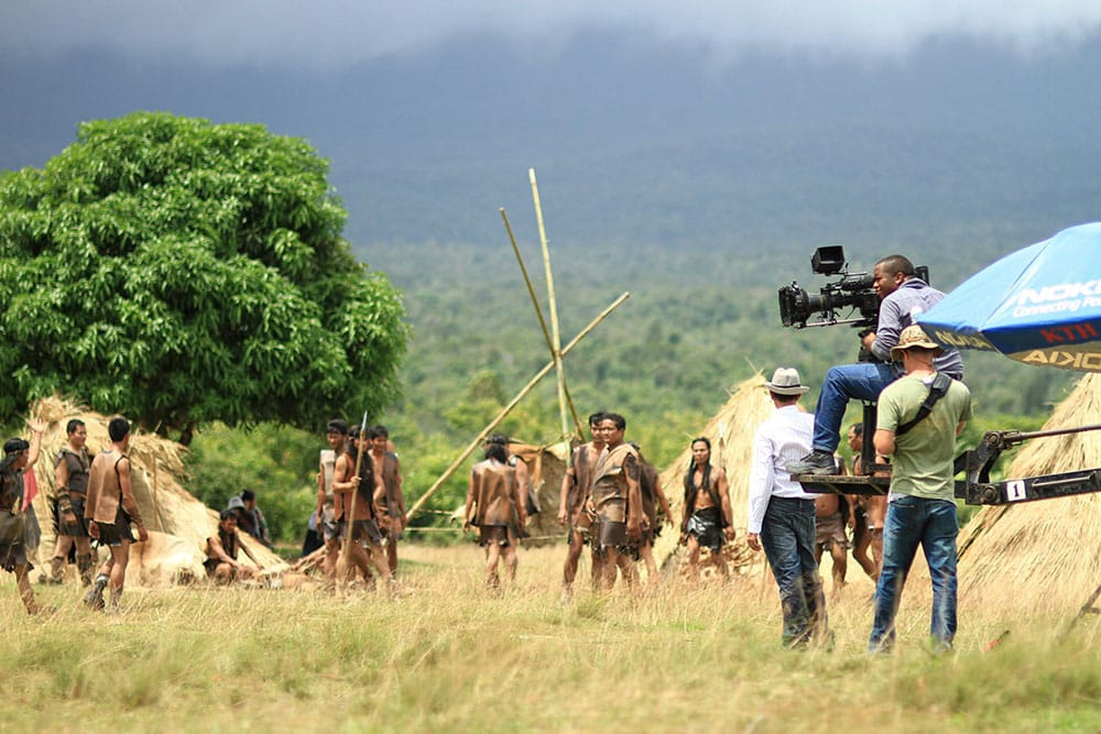 Camera operator sitting on crane filming tribe making straw huts
